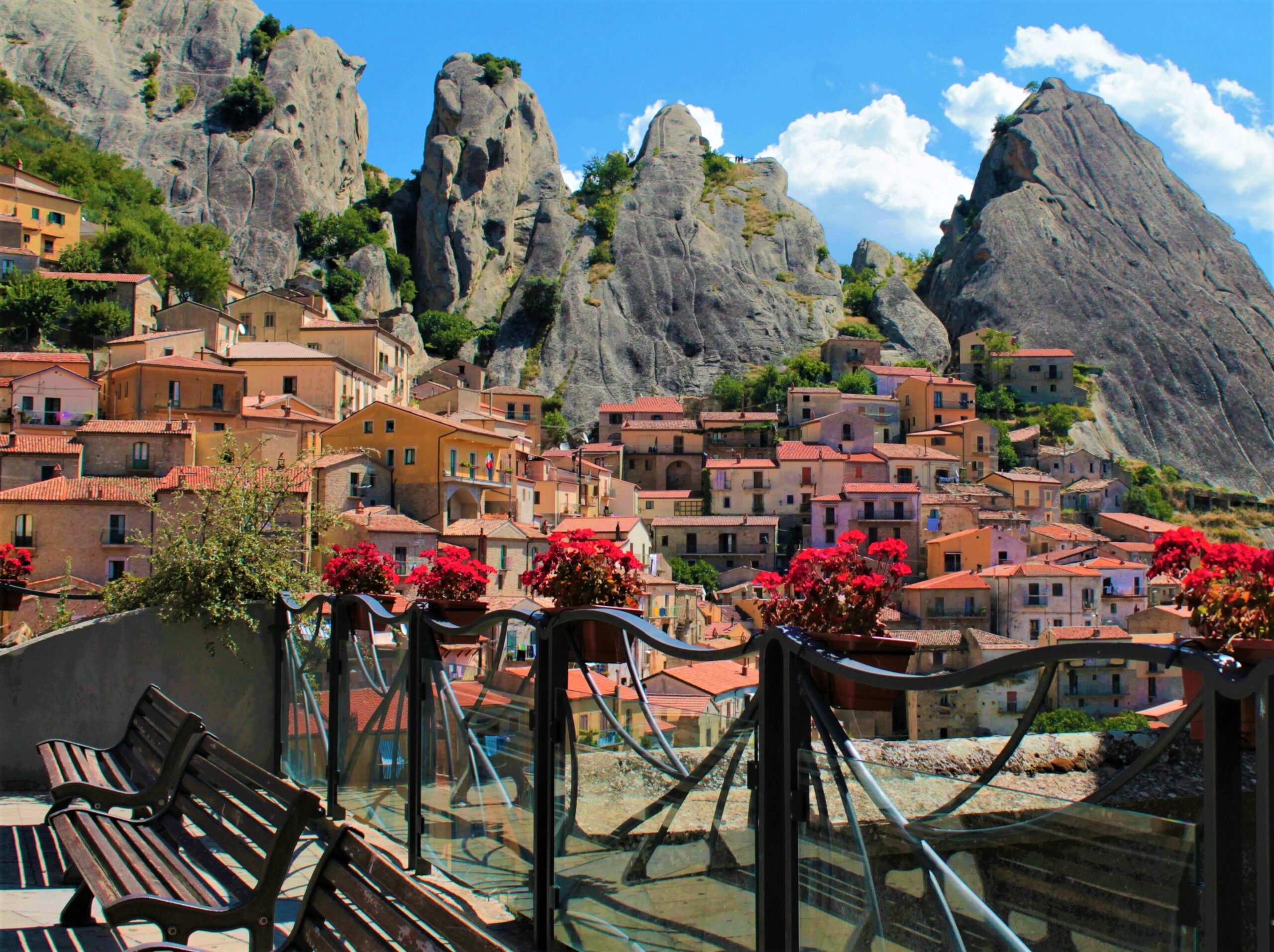 Castelmezzano Vista panoramica di Castelmezzano con sfondo delle Dolomiti lucane. Case rustiche e tetti in terracotta, sullo sfondo di imponenti scogliere grigie. In primo piano si trovano panchine e vivaci fiori rossi, che creano un'atmosfera serena e colorata.