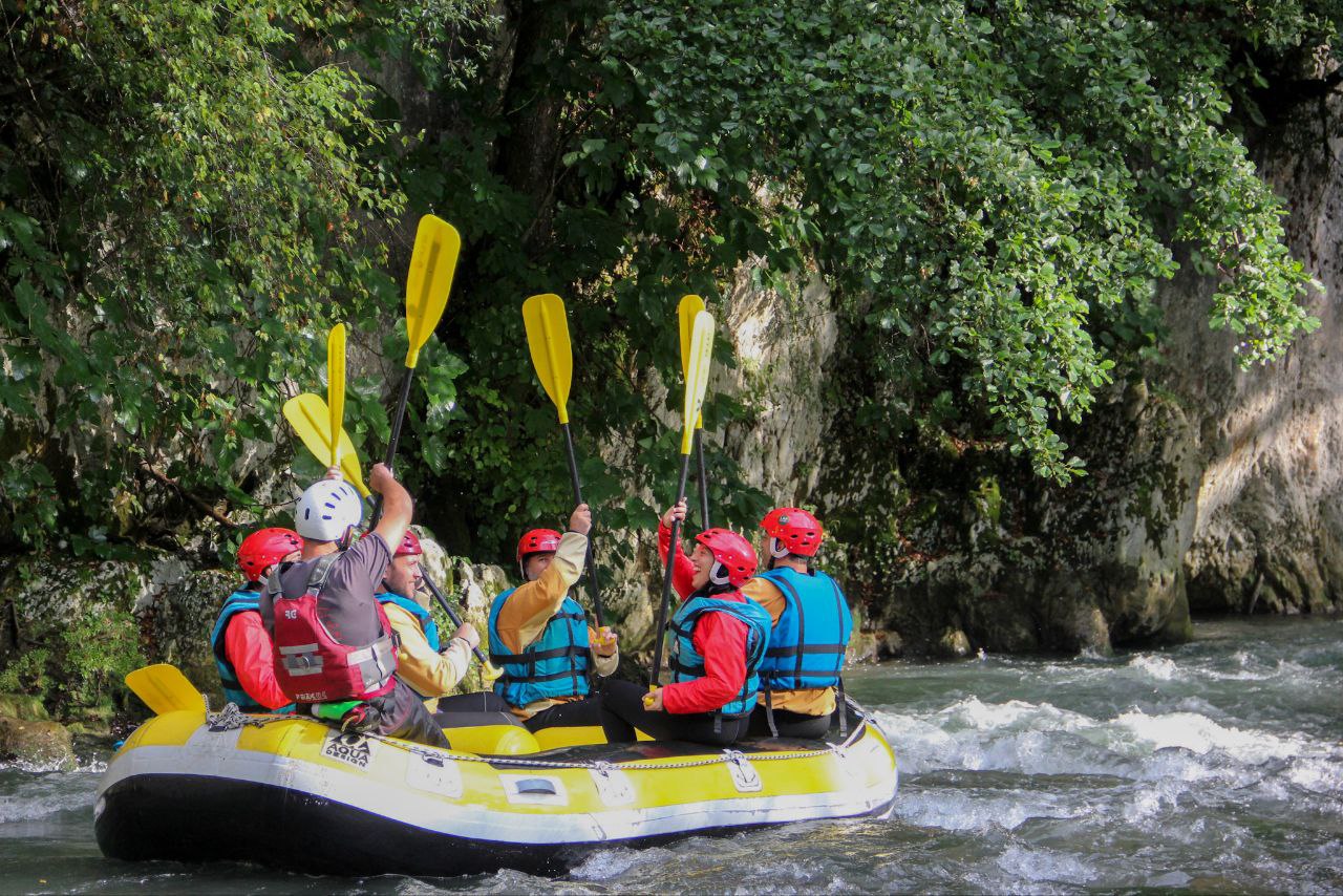 Rafting Un gruppo di persone con caschi e giubbotti di salvataggio fa rafting su un gommone gialla, alzando le pagaie mentre naviga in un fiume circondato dalla foresta, evocando il lavoro di squadra e l'avventura.