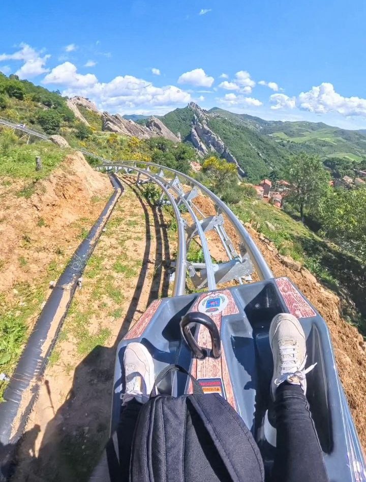Slittovia Vista dalla slittovia delle Dolomiti lucane a Castelmezzano con i piedi nelle scarpe da ginnastica ben visibili, circondato da lussureggianti colline verdi e un cielo azzurro brillante con soffici nuvole.