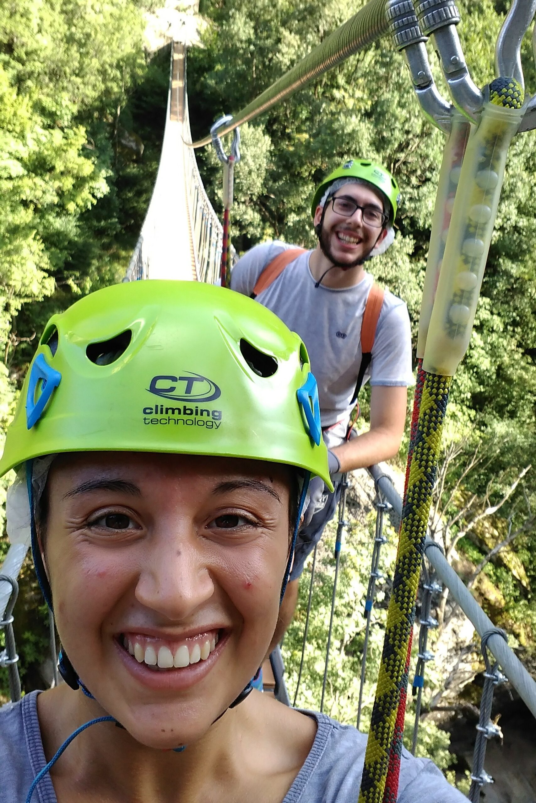 ponte via ferrata 1 Martina e Matteo sorridenti sul ponte sospeso, durante una via ferrata indossando dei caschi da arrampicata di un verde brillante. Lo sfondo mostra una vegetazione lussureggiante, che trasmette emozione e avventura.
