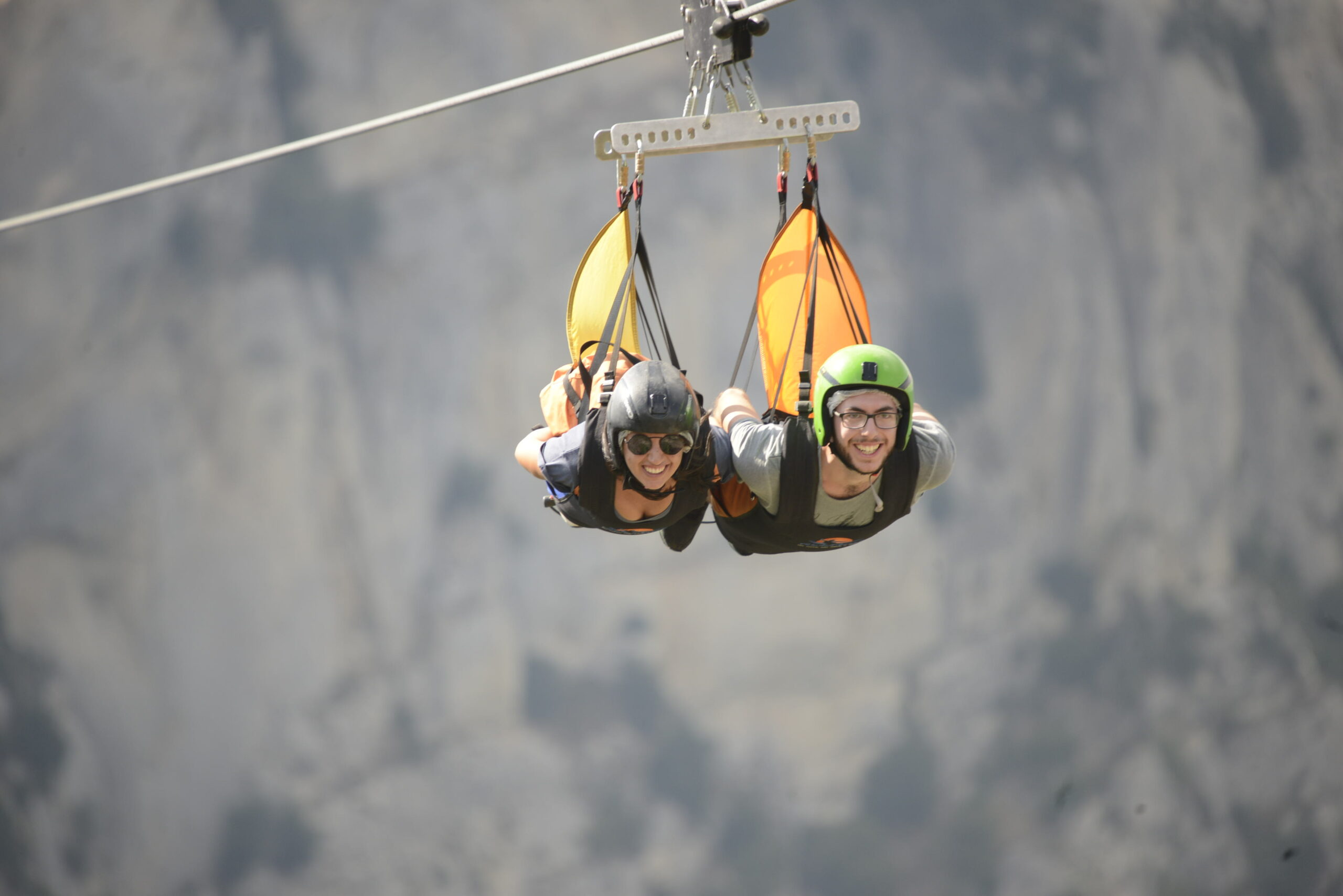 volo dell’angelo Martina e Matteo sorridenti mentre si lanciano in teleferica, una accanto all'altro durante il volo dell'angelo, con casco e attrezzatura, sullo sfondo di un paesaggio naturale sfocato. Avventura ed eccitazione da brivido.