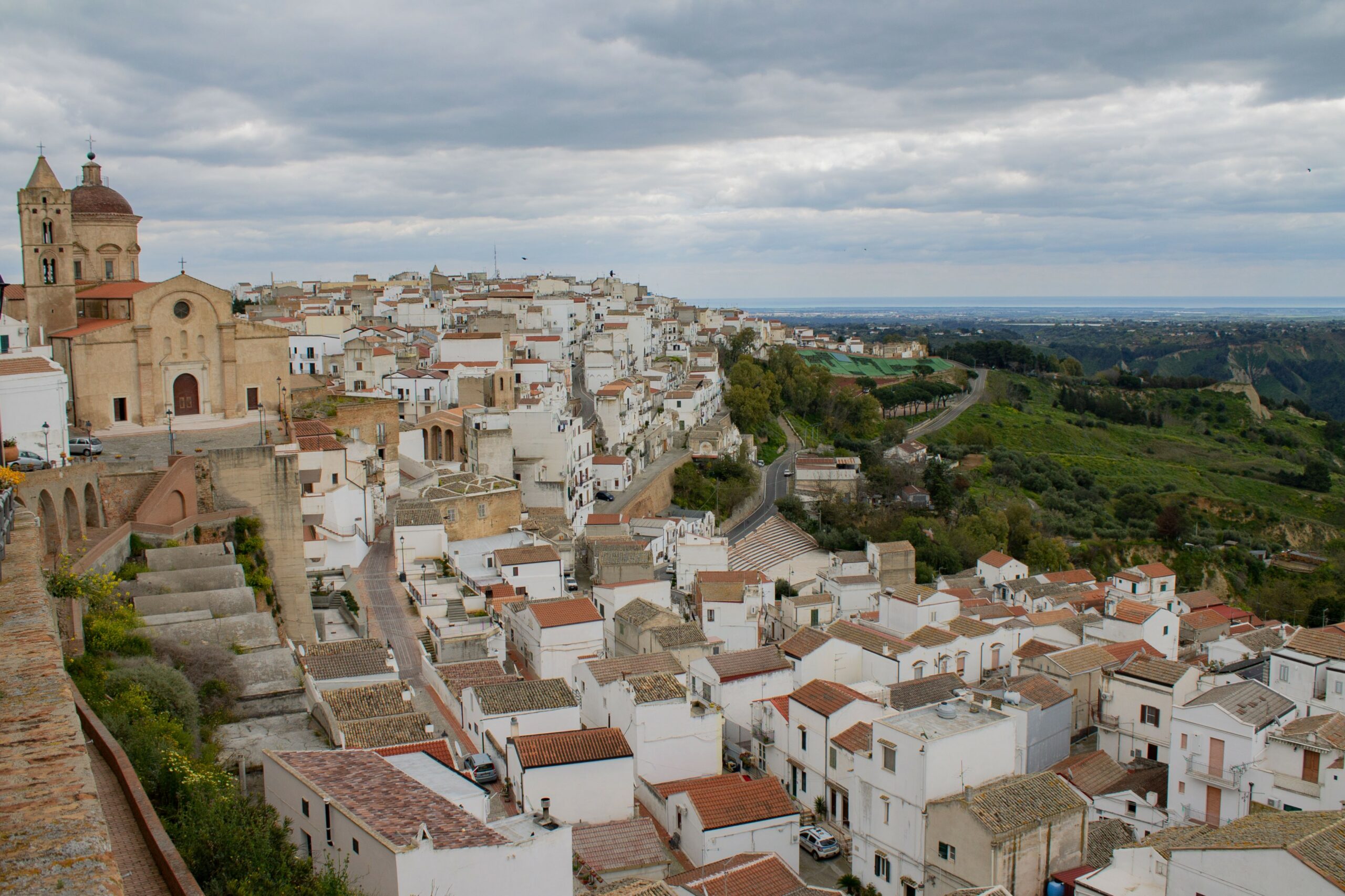 dirupo Vista panoramica sul dirupo di Pisticci, una storica città mediterranea con edifici bianchi e una chiesa. Dolci colline verdi si estendono sotto un cielo coperto. Tranquillo e panoramico.