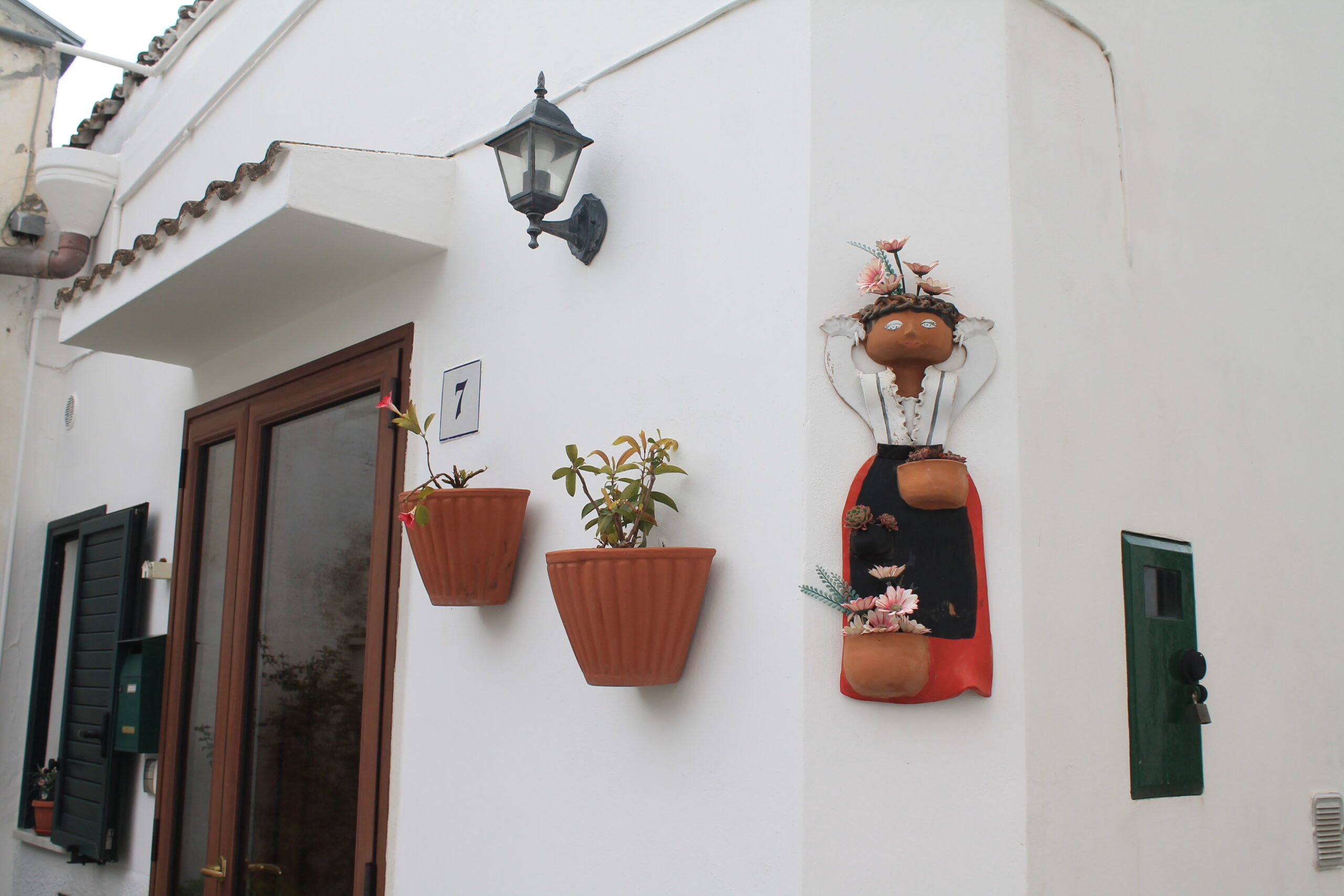 pacchiana 1 Facciata di una casa bianca di Pisticci con una porta marrone, vasi di terracotta appesi al muro con piante, una figura decorativa in ceramica, la pacchiana, e una lampada d'epoca, che trasmettono un'atmosfera pittoresca.