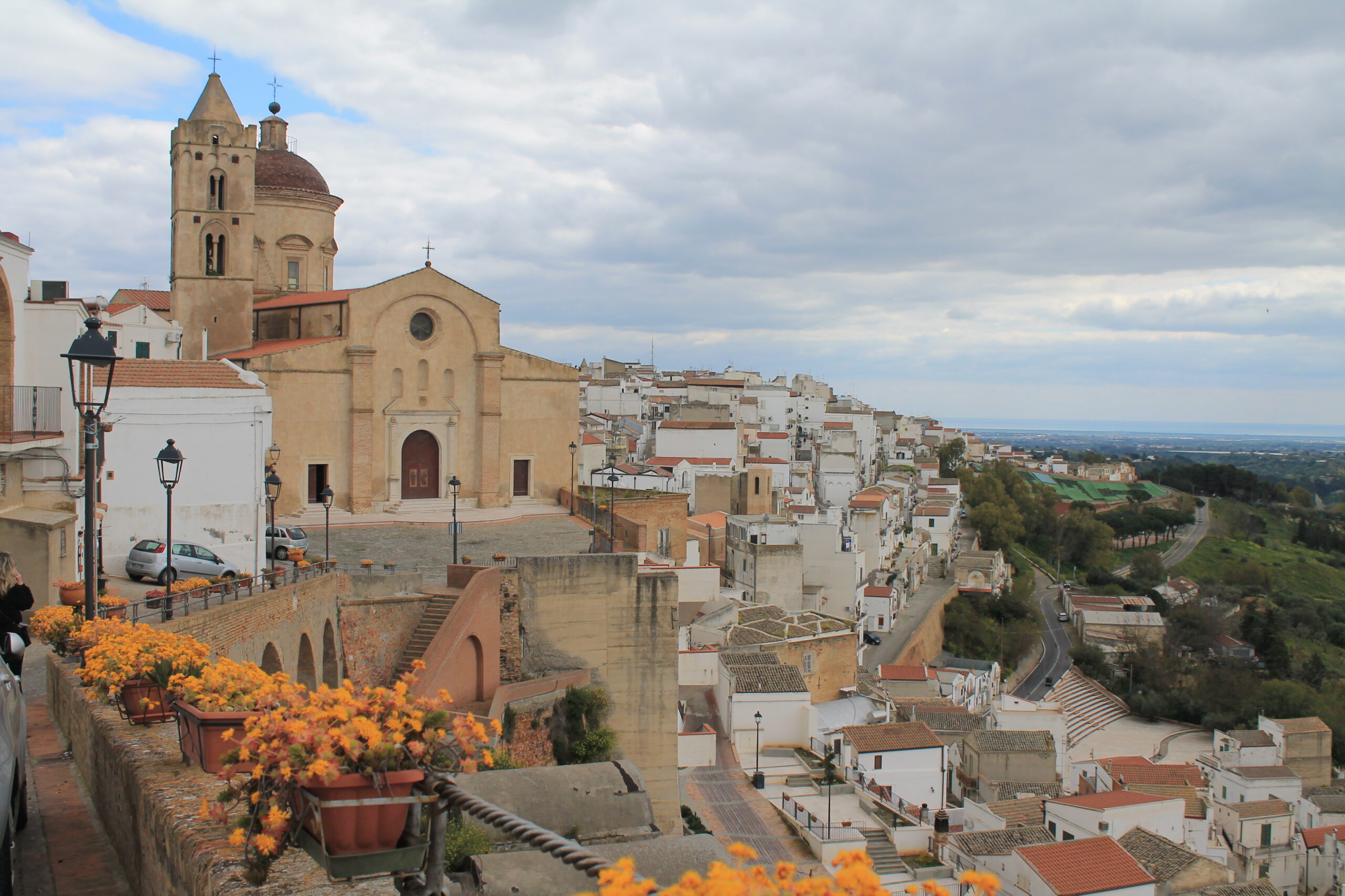 Chiesa storica di Pisticci con torre campanaria in un pittoresco villaggio europeo, circondata da edifici bianchi. Fiori arancioni costeggiano un muro di pietra, sotto un cielo nuvoloso.