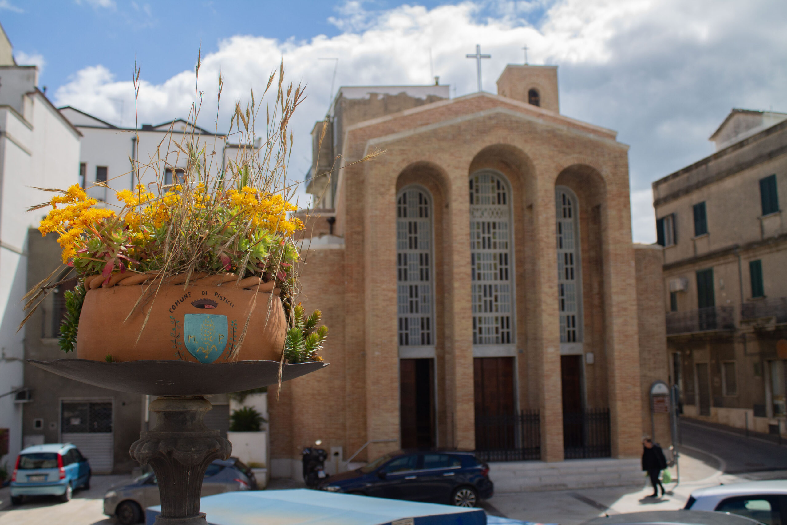 pisticci vaso 1 Fiori gialli ed erba in un vaso con scritto Comune di Pisticci su un piedistallo in primo piano, con una chiesa in mattoni con alte finestre ad arco sullo sfondo. Giornata luminosa e soleggiata.